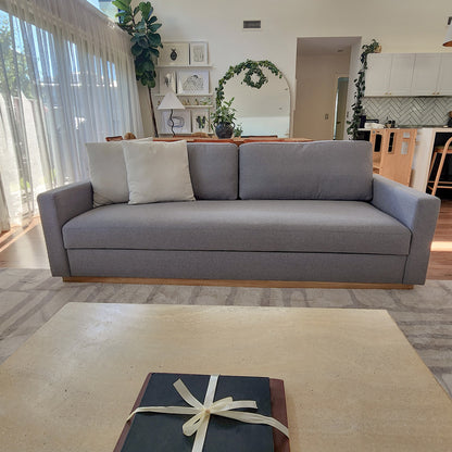 Celeste 3 Seater Hardwood Fabric Sofa Ash on a grey couch with a white pillow, illustrating a modern and stylish design with rubber wood legs for a touch of elegance.