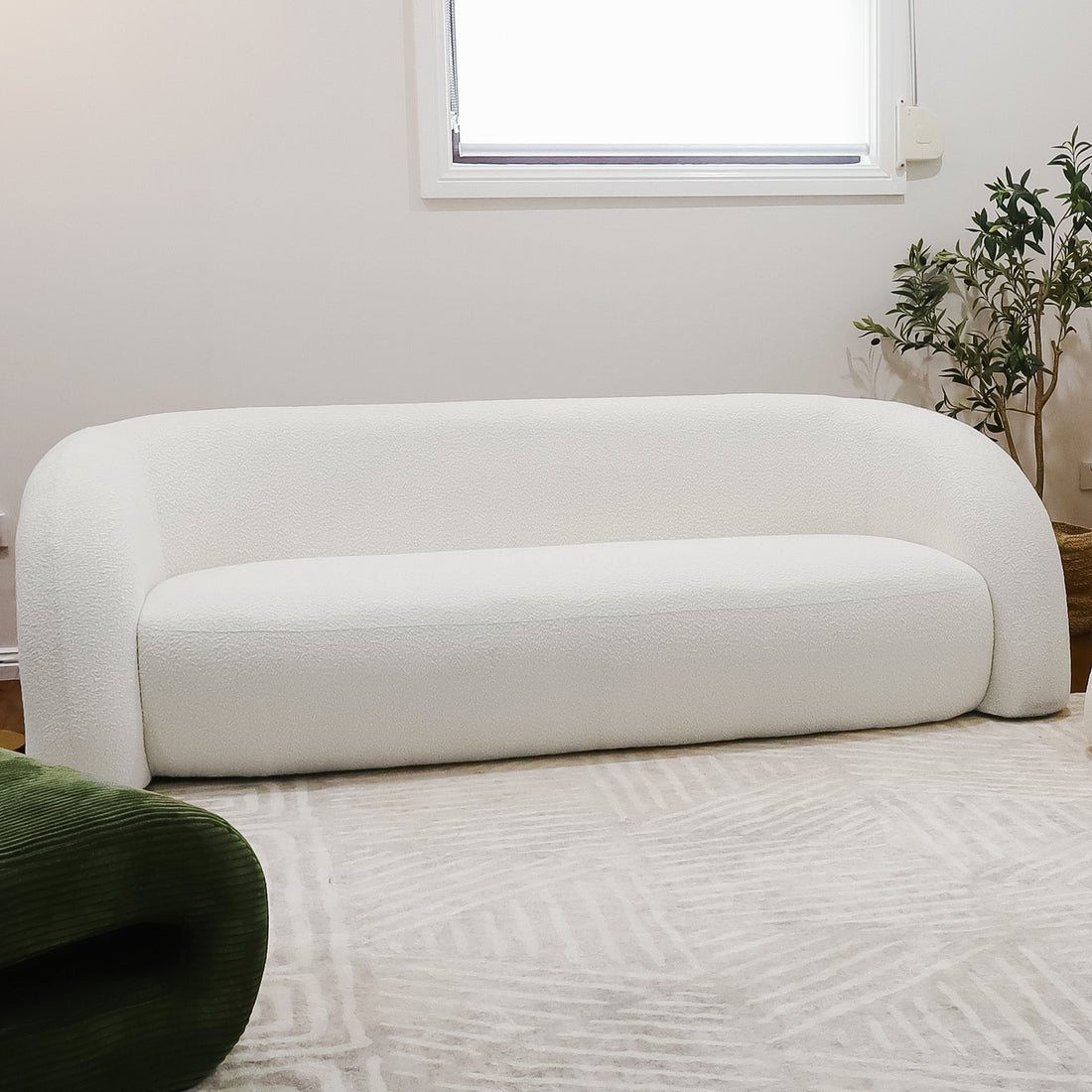 Clioz 3 Seater Curved Boucle Sofa in Eggshell White, featuring rounded arms on a light-patterned rug, with a nearby potted plant and a green fabric piece peeking into the frame.
