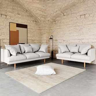 Colton Living Room Package - Cream White features two light-gray sofas with matching throw pillows, a black floor lamp, and an abstract artwork against an exposed-brick wall in a modern space.