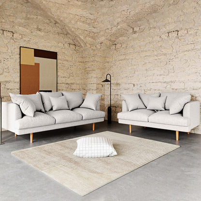 Colton Living Room Package - Cream White features two light-gray sofas with matching throw pillows, a black floor lamp, and an abstract artwork against an exposed-brick wall in a modern space.