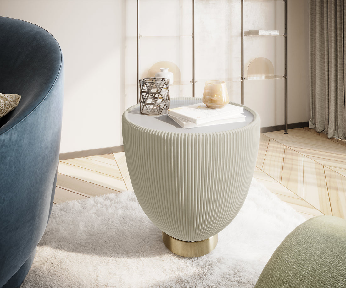 Colton Living Room Package features a pleated-textured stool with a brass base, displayed on a plush rug, accompanied by books and decor, with glimpses of upholstered furniture and a shelving unit nearby.