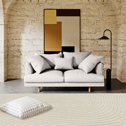 Colton 2 Seater Fabric Sofa with Rug - Cream White: A light-gray two-seat sofa with matching pillows against an exposed stone wall, complemented by a geometric art print and a slender floor lamp.