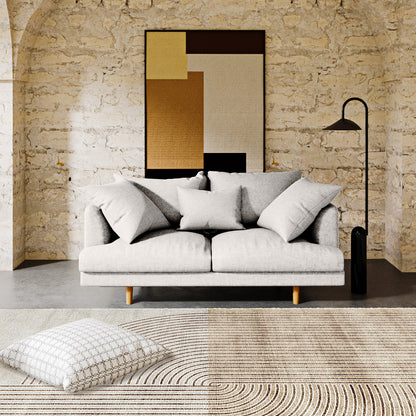 The Colton 2 Seater Fabric Sofa with Rug - Cream White features a minimalist design with a light gray upholstery, three throw pillows, and pairs elegantly with a modern abstract floor rug.