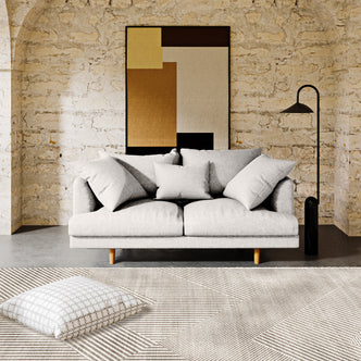 Colton 2 Seater Fabric Sofa with Rug - Cream White; features a light-gray two-seater sofa with throw pillows, framed abstract painting, black floor lamp, and a linear-patterned rug in a loft-style setting.