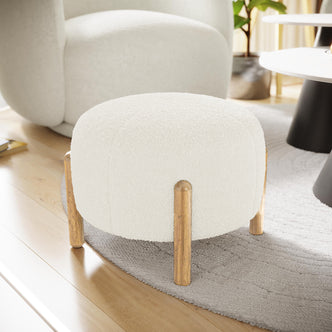 Conchita Boucle Ottoman Stool in soft white boucle fabric, featuring cylindrical wooden legs with a golden finish, sits atop a light gray textured rug in a cozy, minimalist living room setting.