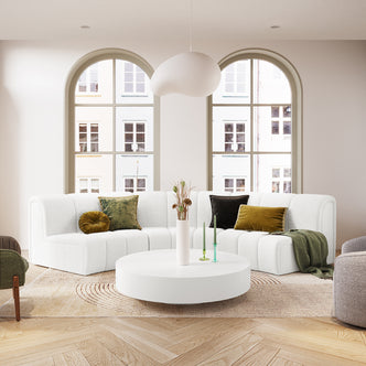 Corina 3 Seater Modular Boucle Sofa in Cream White, L-shaped with muted green and brown accent pillows, green throw, on herringbone wood floor, round coffee table, dried stems, and candles.
