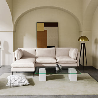 Erling 3 Seater Velvet Sofa with Chaise – Cream, featuring plush velvet upholstery and sleek steel legs, positioned in an elegant indoor setting with modern decor elements like a tripod lamp and abstract painting.