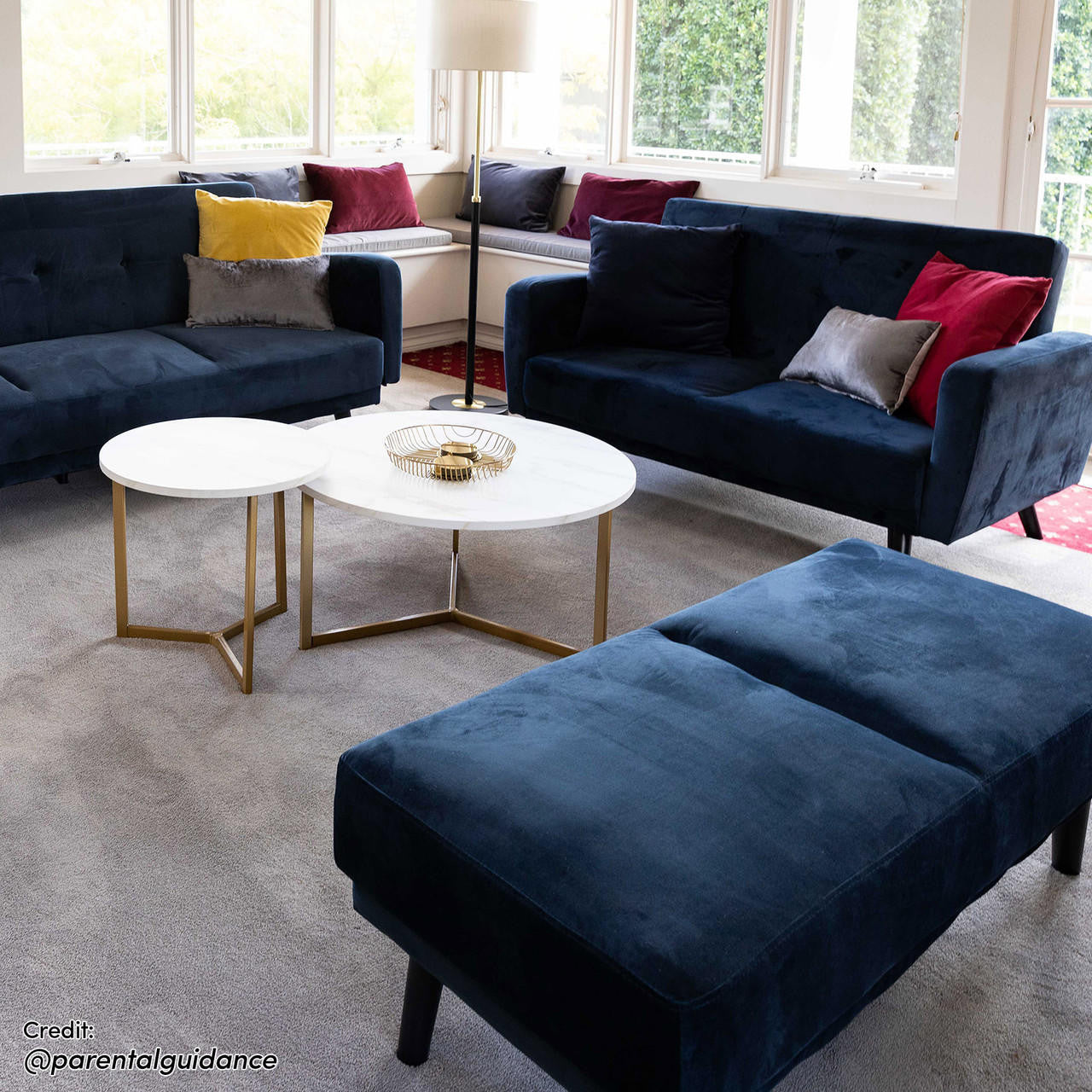 Esben Velvet Fabric Ottoman in Navy Blue with cushioned seat, Scandinavian-style wooden legs, complements navy-blue sofas with colorful pillows in a bright living room featuring nesting tables and large windows.