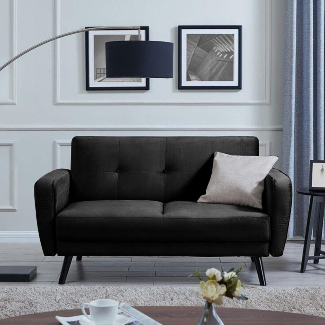 Black Esben 2-Seater Velvet Sofa Bed with button-tufting, featuring a light-gray throw pillow, framed by black-and-white prints, floor lamp, side table, and coffee table with cup, magazine, and flowers.