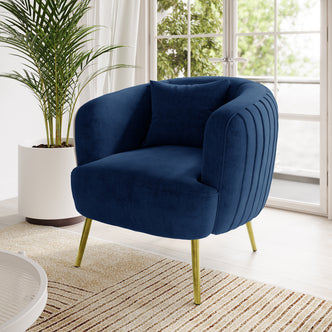 Evelyn Curved Velvet Sofa Accent Chair in Blue with navy velvet upholstery, gold-accented slim legs, and a matching cushion, set against a light wood floor and nearby greenery.