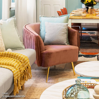 Evelyn Curved Velvet Sofa Accent Chair in Blush with gold accent legs, curved back, and luxurious velvet upholstery, is positioned in a cozy living area next to a light gray sofa and coffee tables.