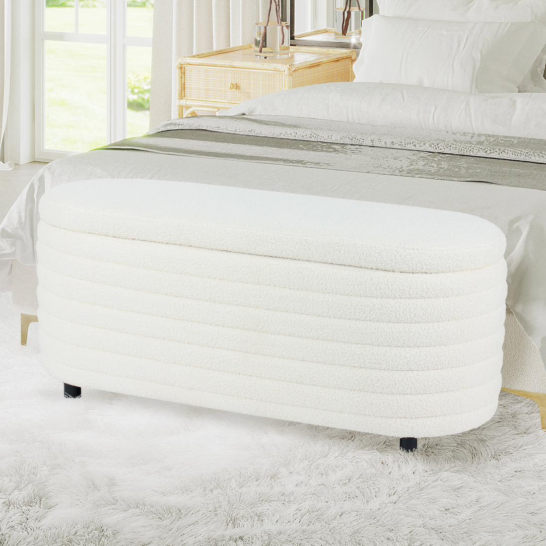 Hartwin 114cm Boucle Ottoman with Storage in Cream White, upholstered in soft bouclé fabric, with short black feet, situated in a bright bedroom, in front of a bed with white linens.