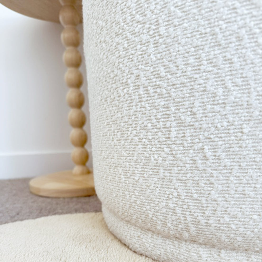 Close-up of the Hiboa Bouclé Ottoman Stool in Eggshell White, featuring textured bouclé fabric and a round design. It rests on a circular rug with a wooden side table visible in the background.