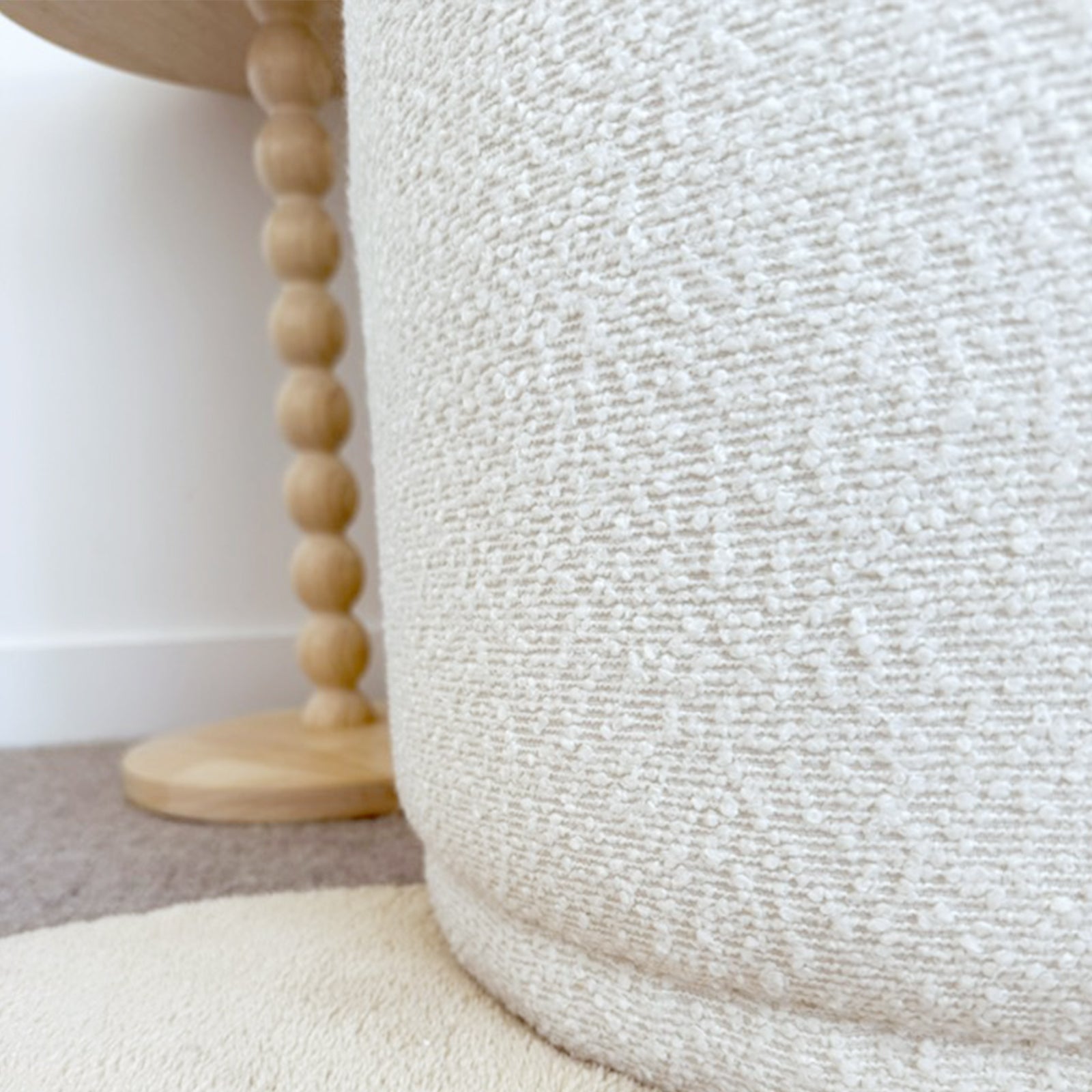 Close-up of the Hiboa Bouclé Ottoman Stool in Eggshell White, featuring textured bouclé fabric and a round design. It rests on a circular rug with a wooden side table visible in the background.