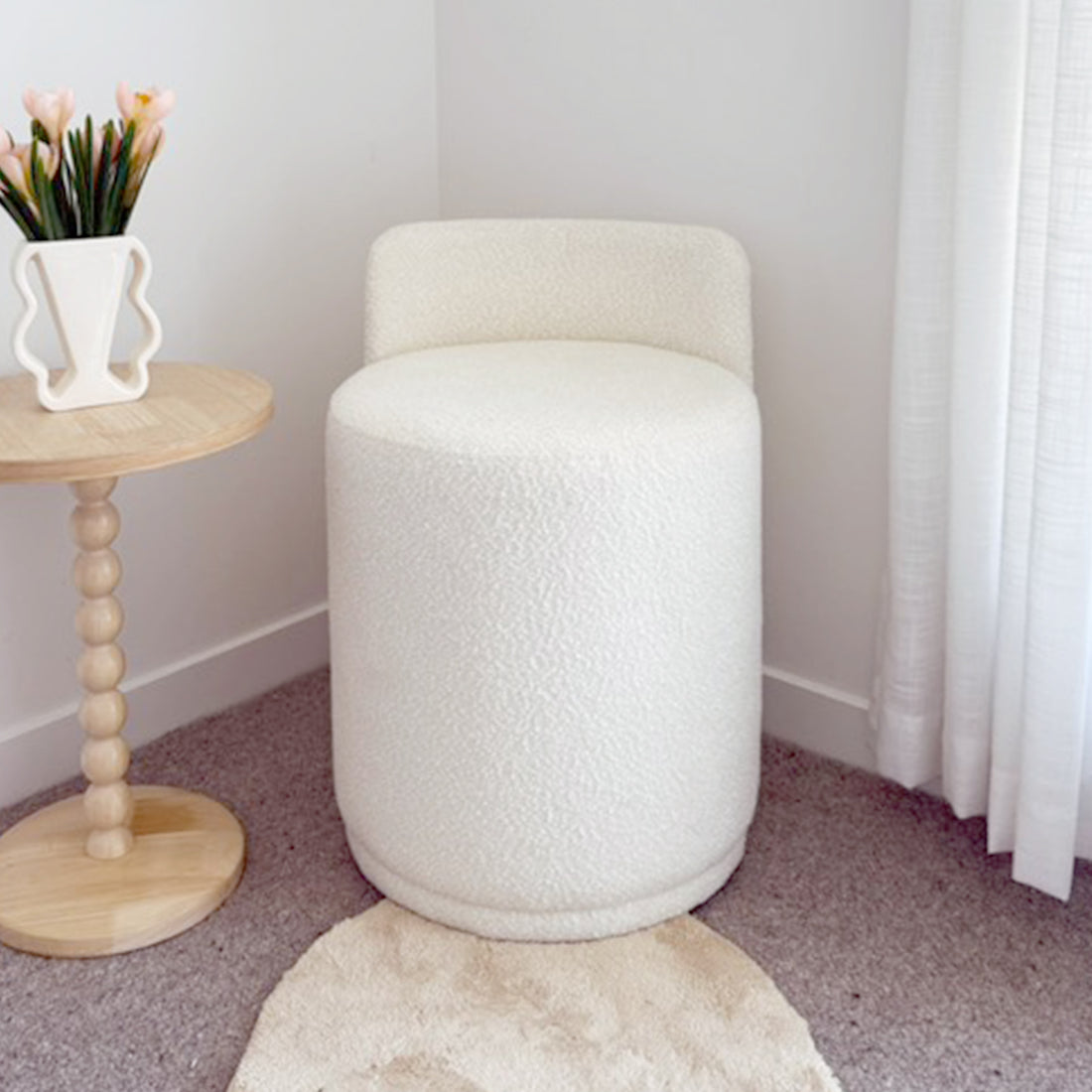 Hiboa Bouclé Ottoman Stool in Eggshell White, cylindrical with a tiny backrest, sits on a neutral-tone carpet, near a wooden side table and light curtains, showcasing its cozy, textured upholstery.