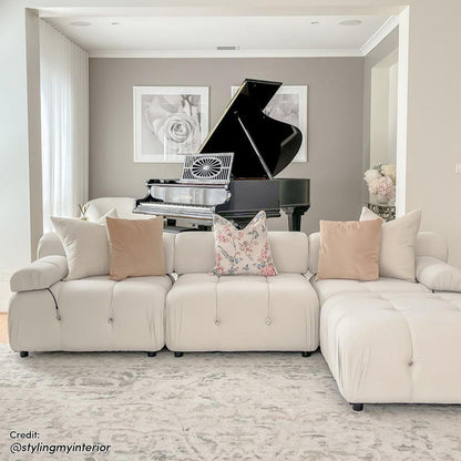 Keila 3 Seater Modular Tufted Sofa with Ottoman in Cream, positioned in a serene living room with a white sectional design, surrounded by elegant decor including a grand piano and botanical prints.