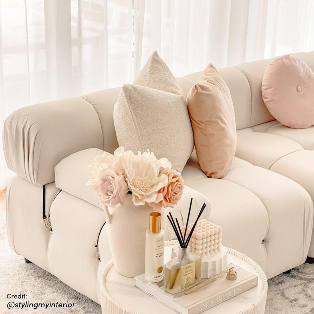 Keila 3 Seater Modular Tufted Sofa with Ottoman in Cream, featuring three throw pillows, a side table with decor items, and sheer drapes in the background, showcasing a modern living room setting.