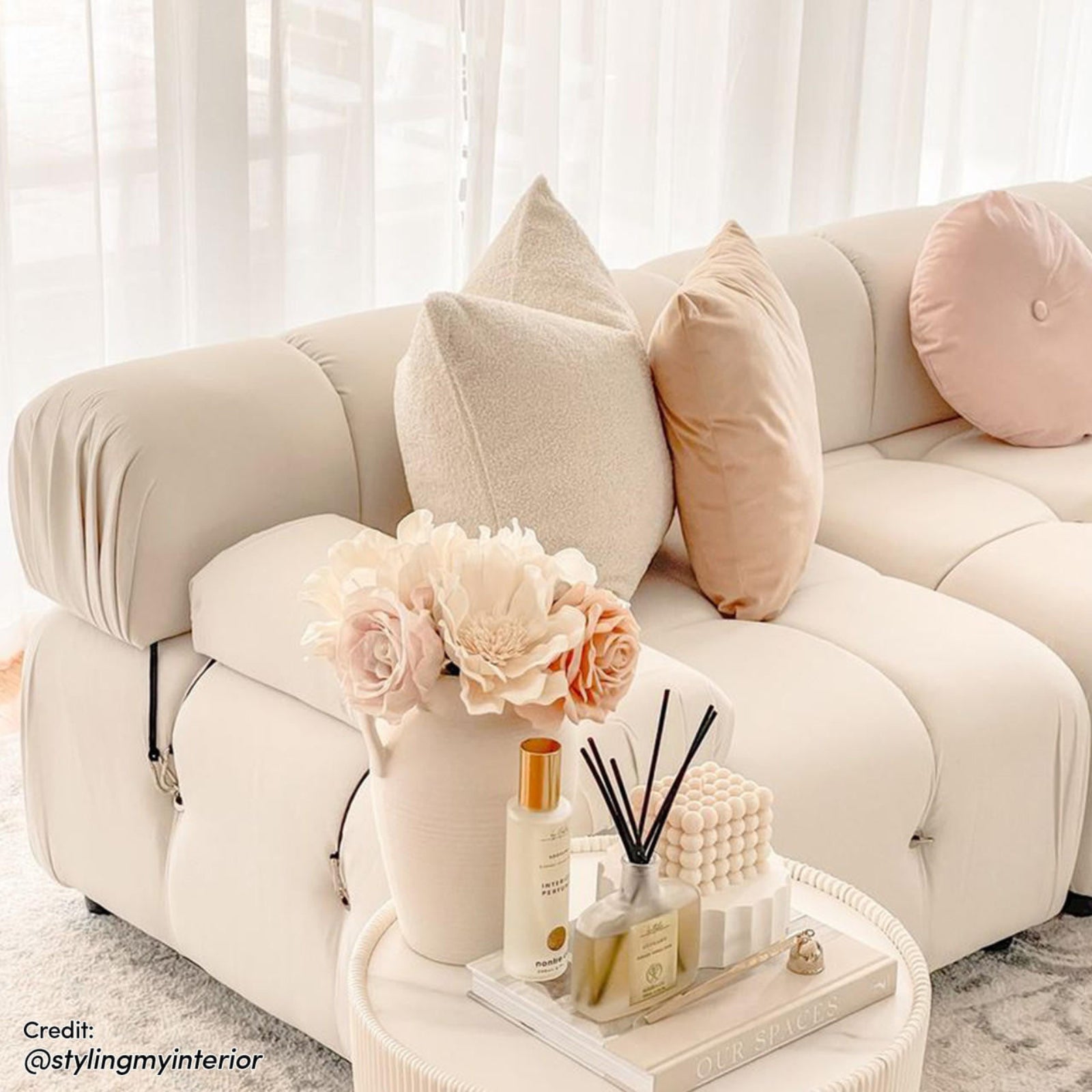 Keila 3 Seater Modular Tufted Sofa with Ottoman in Cream, featuring three throw pillows, a side table with decor items, and sheer drapes in the background, showcasing a modern living room setting.