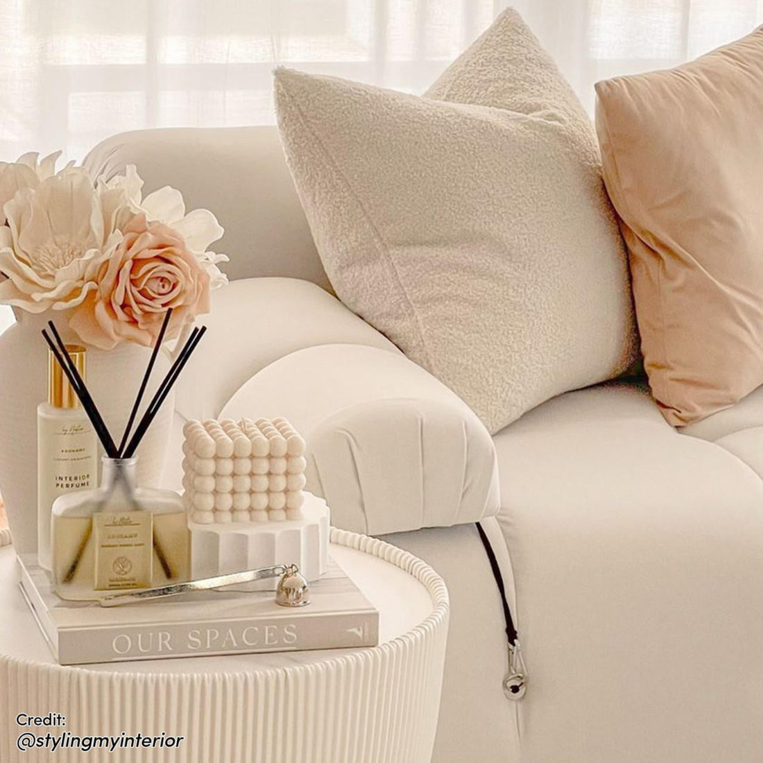 Keila 3 Seater Modular Tufted Sofa with Ottoman in Cream, featuring plush velvet upholstery, two throw pillows, and a fluted-sided side table with a decorative arrangement beside it.