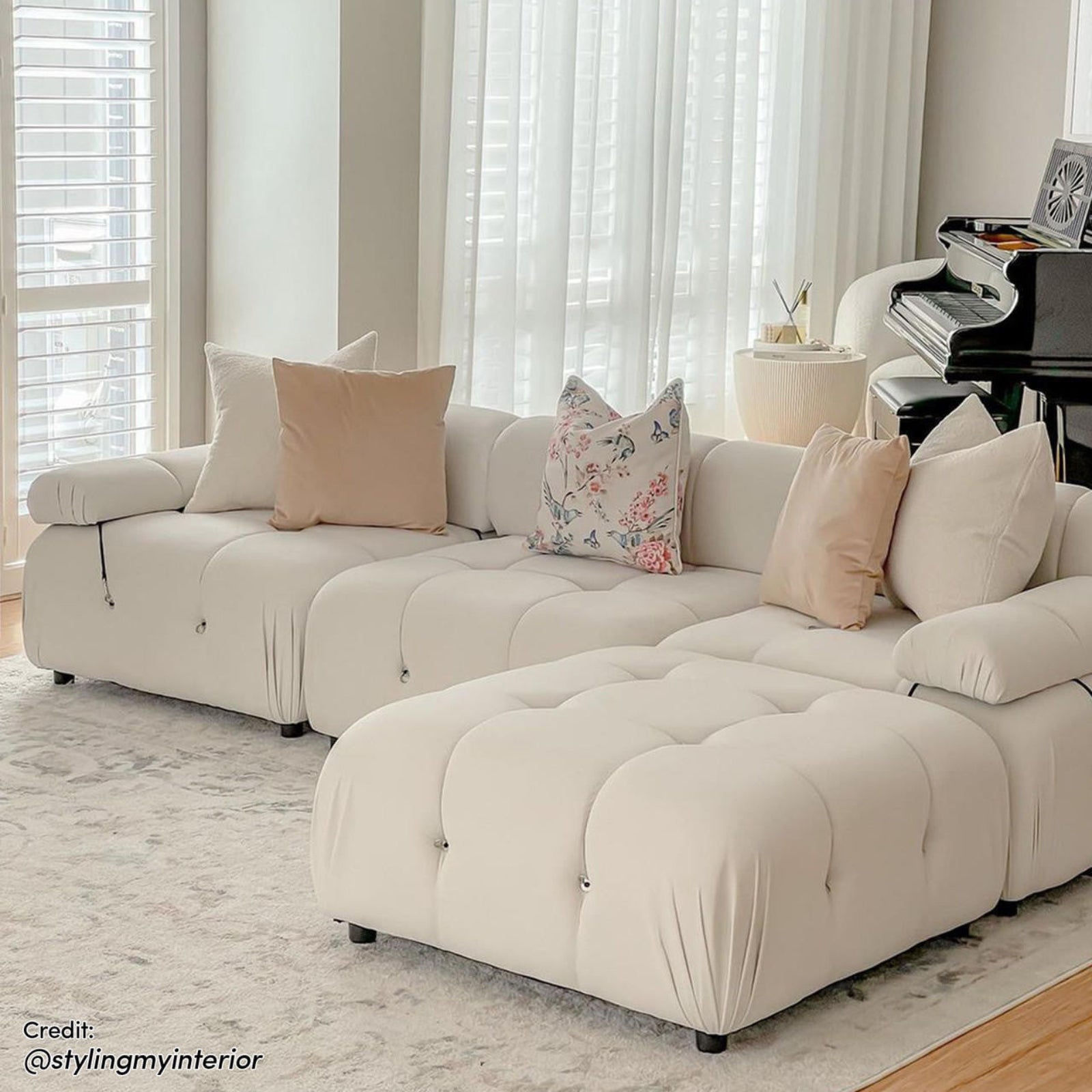 Keila 3 Seater Modular Tufted Sofa with Ottoman in Cream displayed in a bright living area, featuring blush pillows, floral-butterfly print, and complemented by a small round side table and upright piano.