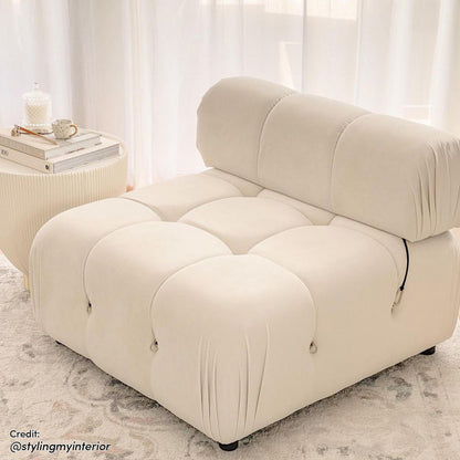 Keila Modular Tufted Single Sofa in Cream Velvet features oversized, quilt-style tufted blocks, creating a plush, sculptural look. Paired with a small side table holding books and a mug.