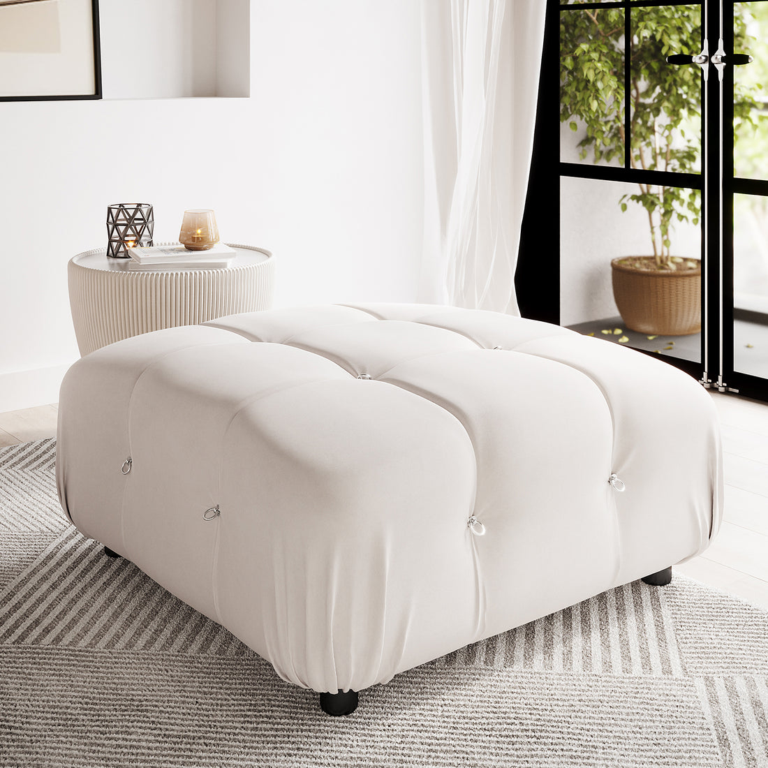 Keila Modular Tufted Velvet Ottoman in Soft Pink, with elegant tufting, sits in a minimal interior corner on a striped rug, accompanied by a ribbed side table holding decorative items.