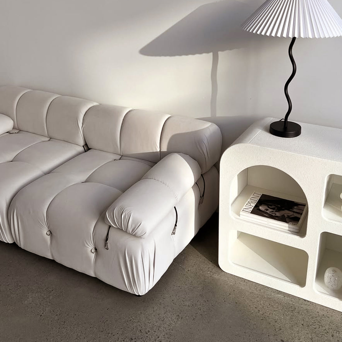 Keila 2 Seater Modular Tufted Sofa Set in Cream Velvet with tufted cushions and exposed zippers, next to a textured side unit holding magazines and a lamp, on polished concrete floor.