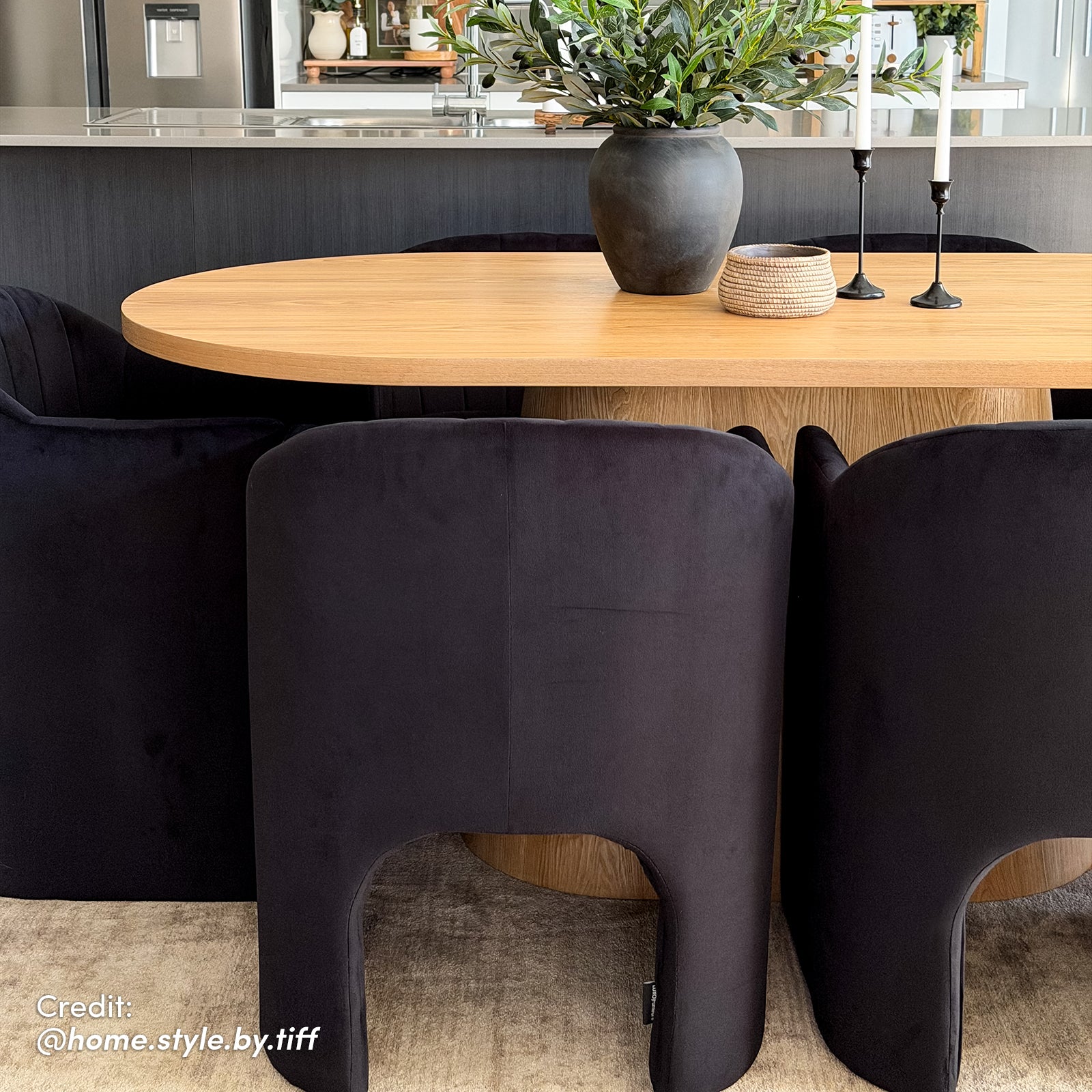 Liora Velvet Curved Accent Armchair in Black featured in a modern dining setup, surrounded by black chairs, oval wood table, and decor elements like a gray vase and candle holders.