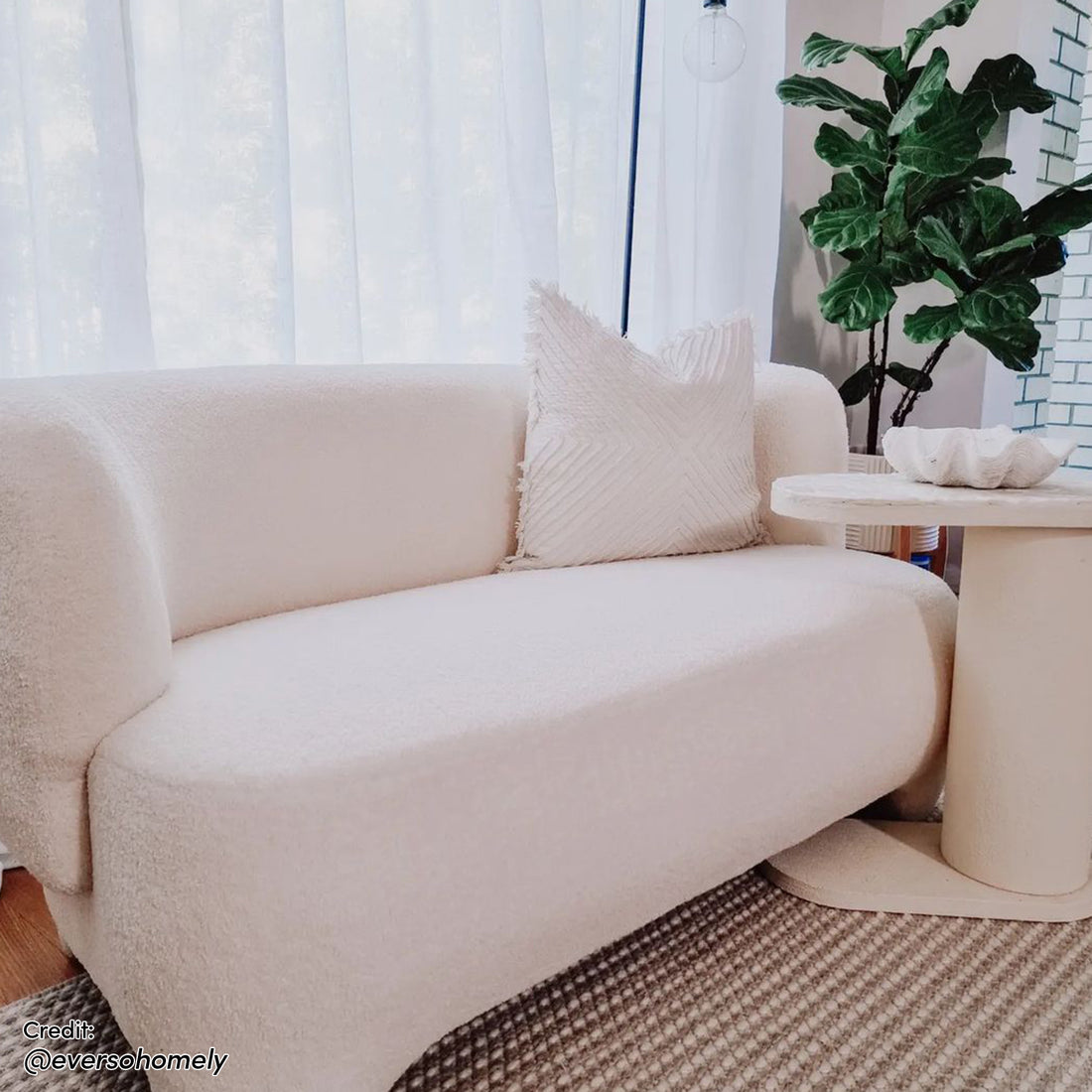 Cream-colored, softly curved Massie 2-Seater Boucle Sofa with an off-white textured throw pillow, complemented by a matching side table, sculptural bowl, and natural-fiber rug for a modern, cozy aesthetic.
