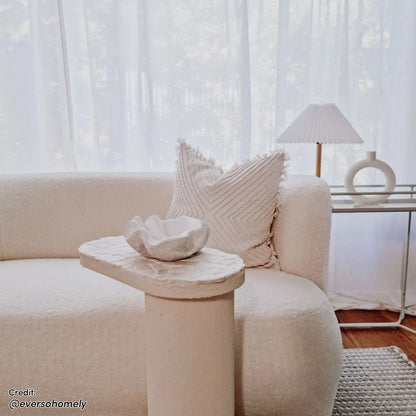 Massie 2-Seater Boucle Sofa - Cream White, plush and textured, with a rounded design, resting in a cozy indoor setting featuring a white pillow and a lamp by a window.