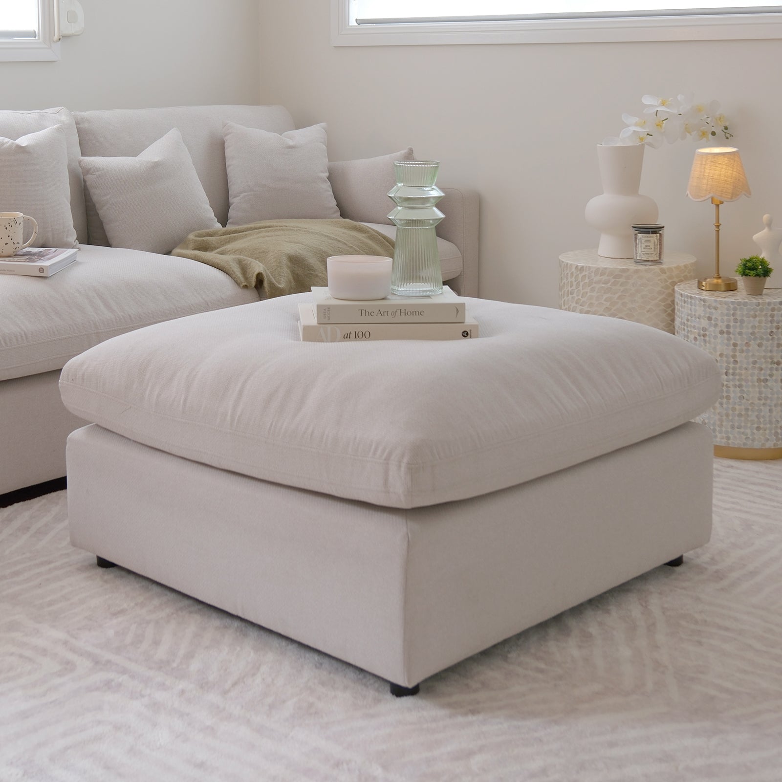 Millie Fabric Ottoman Modular Sofa in warm light grey on a couch, showcasing its plush, deep seating and sturdy timber frame, complemented by a white pillow and lamp in an inviting interior setting.