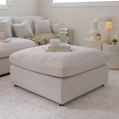 Millie Fabric Ottoman Modular Sofa in warm light grey on a couch, showcasing its plush, deep seating and sturdy timber frame, complemented by a white pillow and lamp in an inviting interior setting.