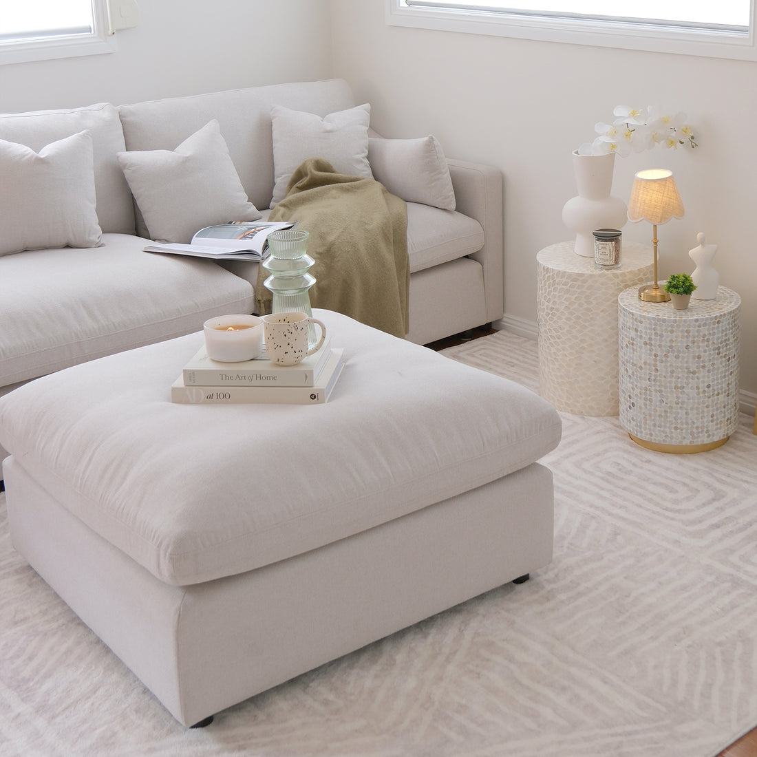 Millie Fabric Ottoman Modular Sofa in warm light grey, featuring clean lines, a plush surface, and a sturdy frame. Positioned with a white pillow, candle, and books, exuding a cozy interior vibe.