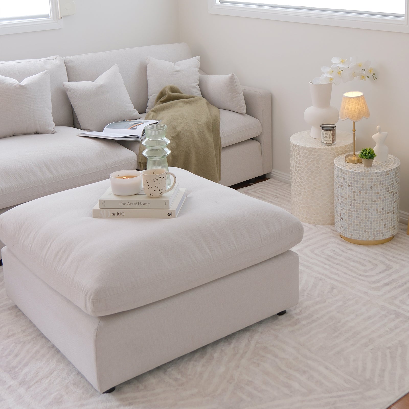 Millie Fabric Ottoman Modular Sofa in warm light grey, featuring clean lines, a plush surface, and a sturdy frame. Positioned with a white pillow, candle, and books, exuding a cozy interior vibe.