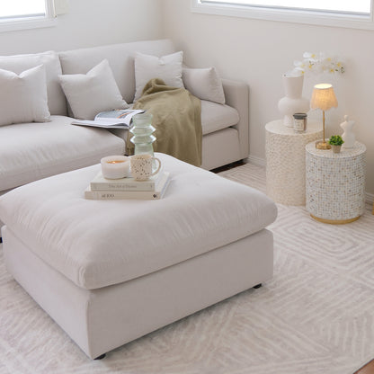 Millie Fabric Ottoman Modular Sofa in warm light grey, featuring clean lines, a plush surface, and a sturdy frame. Positioned with a white pillow, candle, and books, exuding a cozy interior vibe.