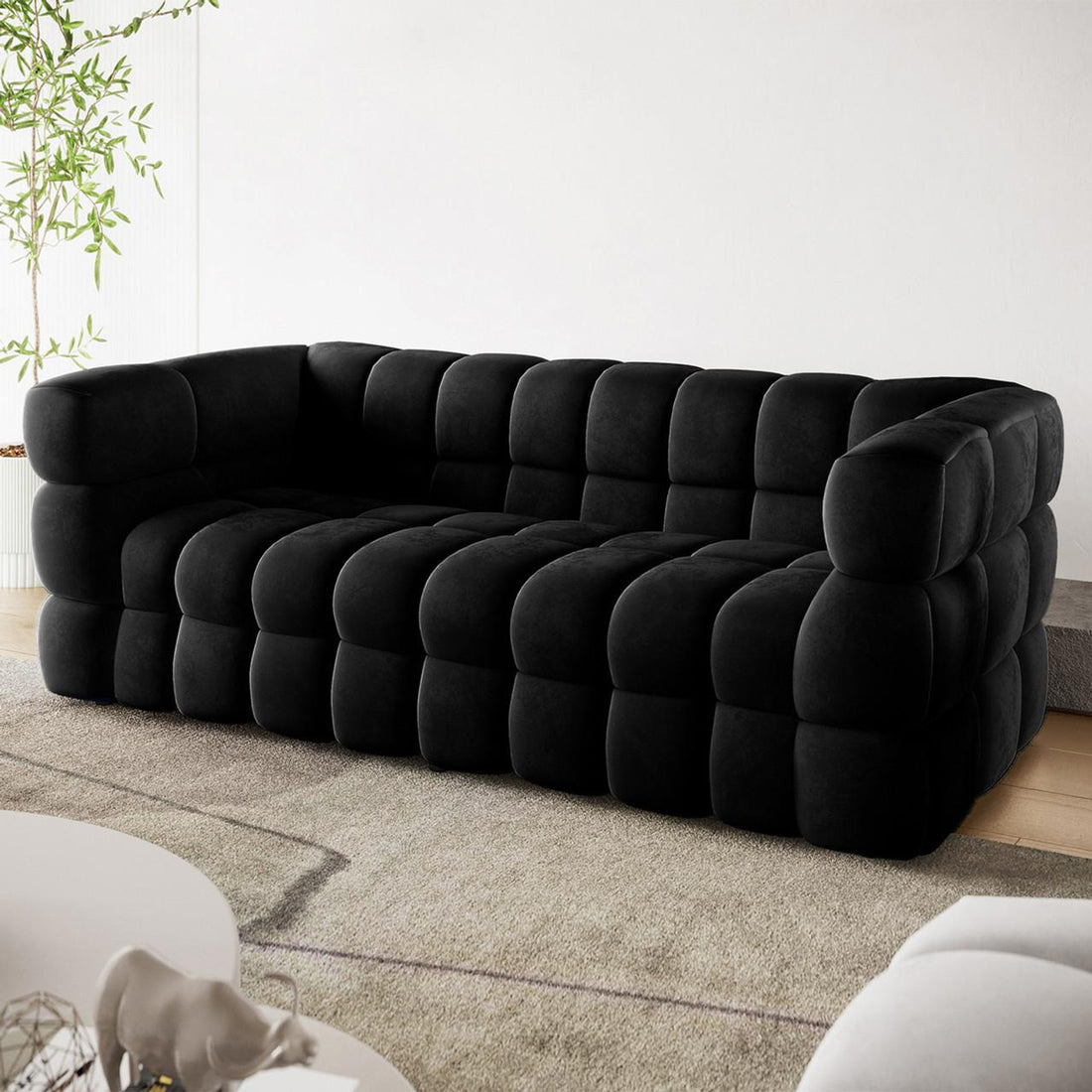 The Norah 3 Seater Velvet Sofa in Black features oversized padded squares creating a modern tufted design, placed centrally in a chic living room setup with a white coffee table and potted plant.