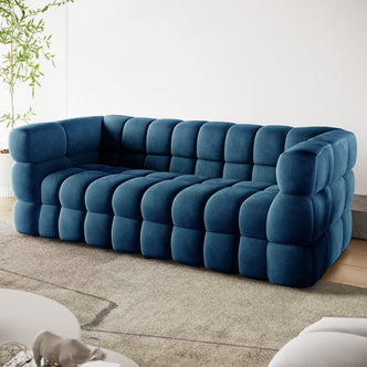 Norah 3 Seater Velvet Sofa in Blue, featuring a chunky grid-tufted design with large square cushions. Set in a modern living room, it exudes comfort and elegance in a minimalist setting.