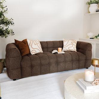 The Norah 3 Seater Boucle Sofa in Brown, tufted and chocolate-brown, is adorned with cream and rust pillows, complemented by a round coffee table and stylish living room decor.