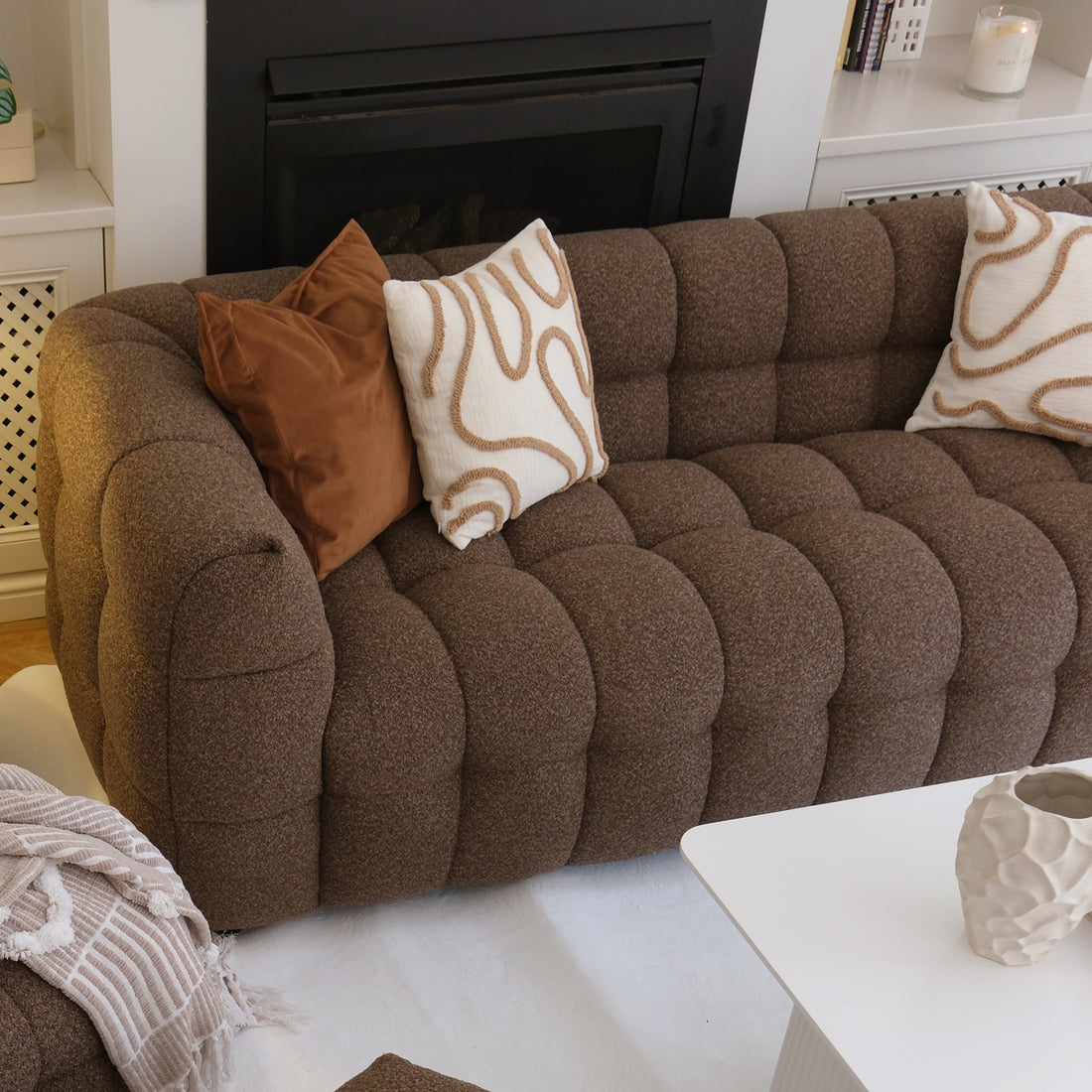 Norah 3 Seater Boucle Sofa in Brown, tufted and cozy with decorative pillows and a beige throw, anchored by a white coffee table and surrounded by a warm, inviting living room setting.
