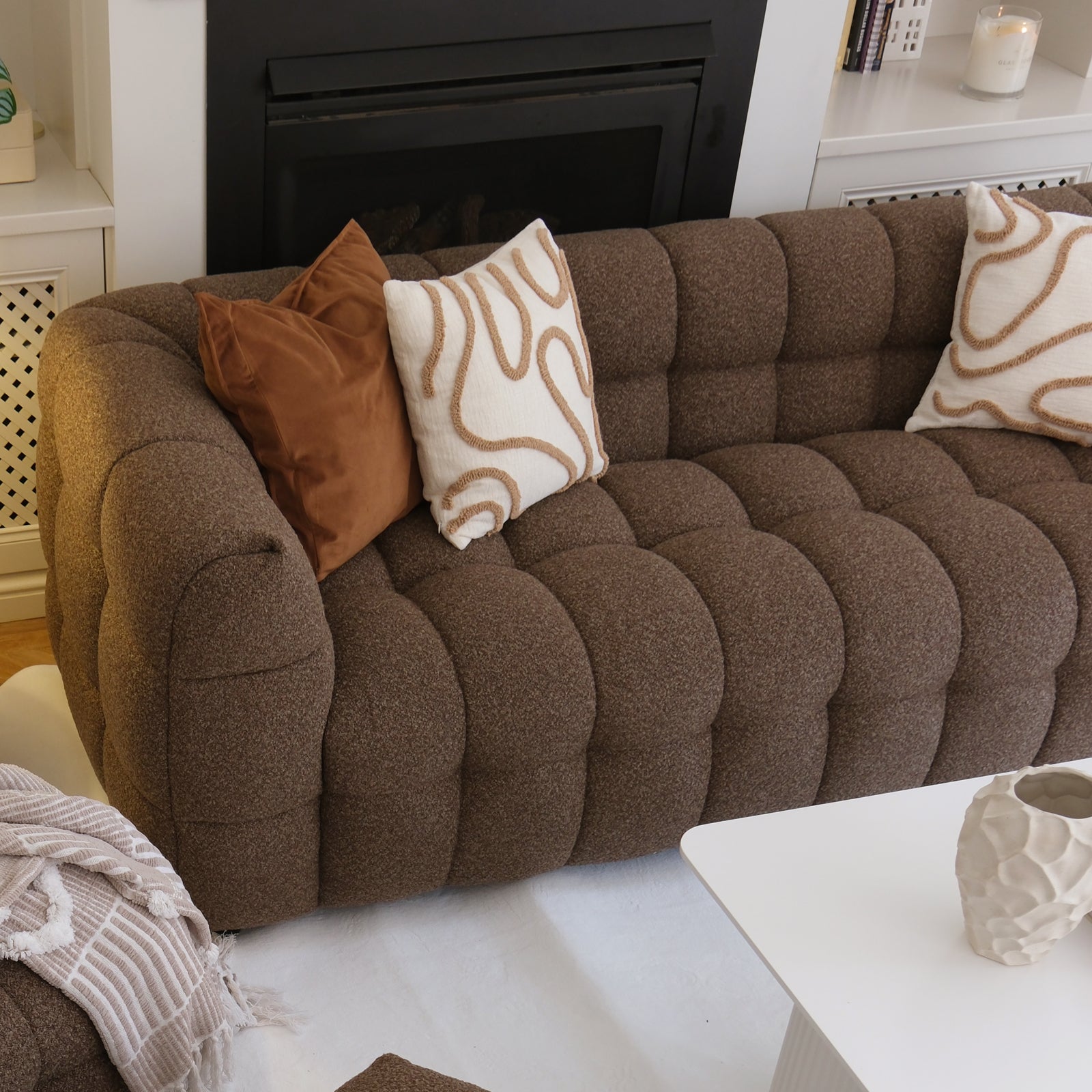 Norah 3 Seater Boucle Sofa in Brown, tufted and cozy with decorative pillows and a beige throw, anchored by a white coffee table and surrounded by a warm, inviting living room setting.