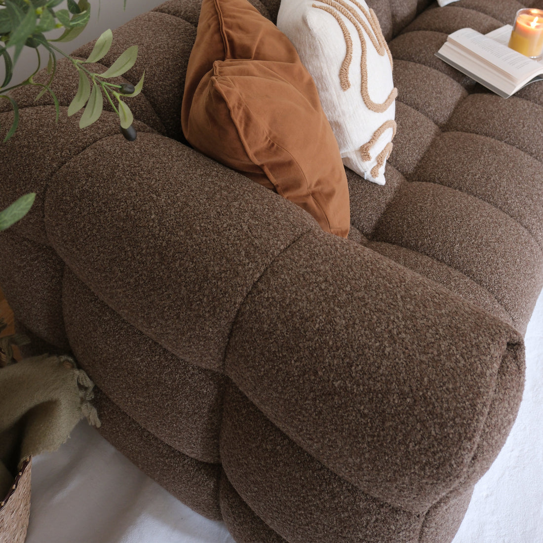 Norah 3 Seater Boucle Sofa in Brown, featuring plush chocolate-brown fabric, two decorative throw pillows, a lit candle, open book, and peeking houseplant, creating a cozy, inviting living-room scene.
