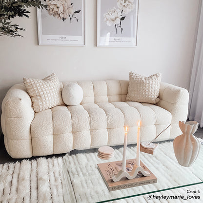 Norah 3 Seater Boucle Sofa in Cream White, featuring tufted square sections, two textured pillows, and a round cushion, placed in a serene, neutral-toned living room setting with modern decor elements.