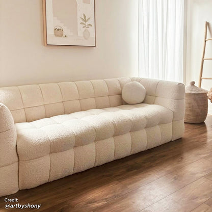 Norah 3 Seater Boucle Sofa in Cream White features a plush, tufted design with rounded corners and a matching bolster pillow, set on hardwood floors in a cozy, modern-minimalist living space.
