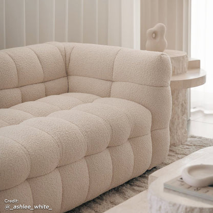 Norah 3 Seater Boucle Sofa in Cream White features softly curved lines, tufted cushions, and boucle fabric, complemented by a minimalist setup with rounded tables and soft textures.