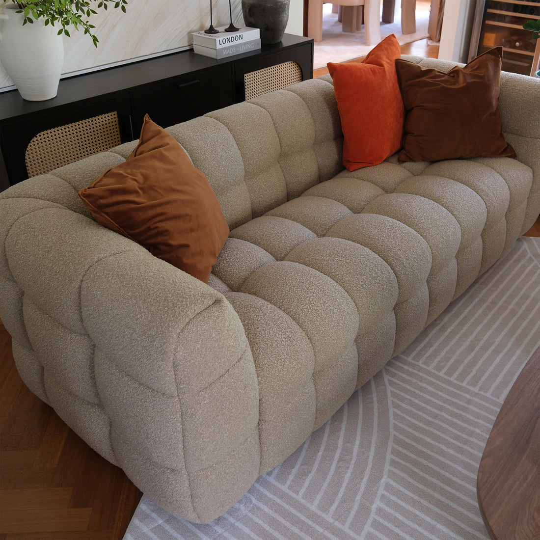 Norah 3 Seater Boucle Sofa in Warm Beige, featuring rounded edges and tufted design with nubby texture, accented by three oversized throw pillows, displayed in a cozy living room setting.