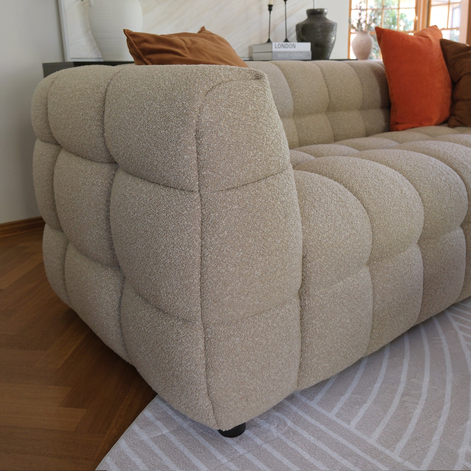 Norah 3 Seater Boucle Sofa in Warm Beige, showcasing its quilted bubble cushions in cream-beige boucle fabric, accented with rust pillows, on a curved-patterned rug over hardwood flooring.