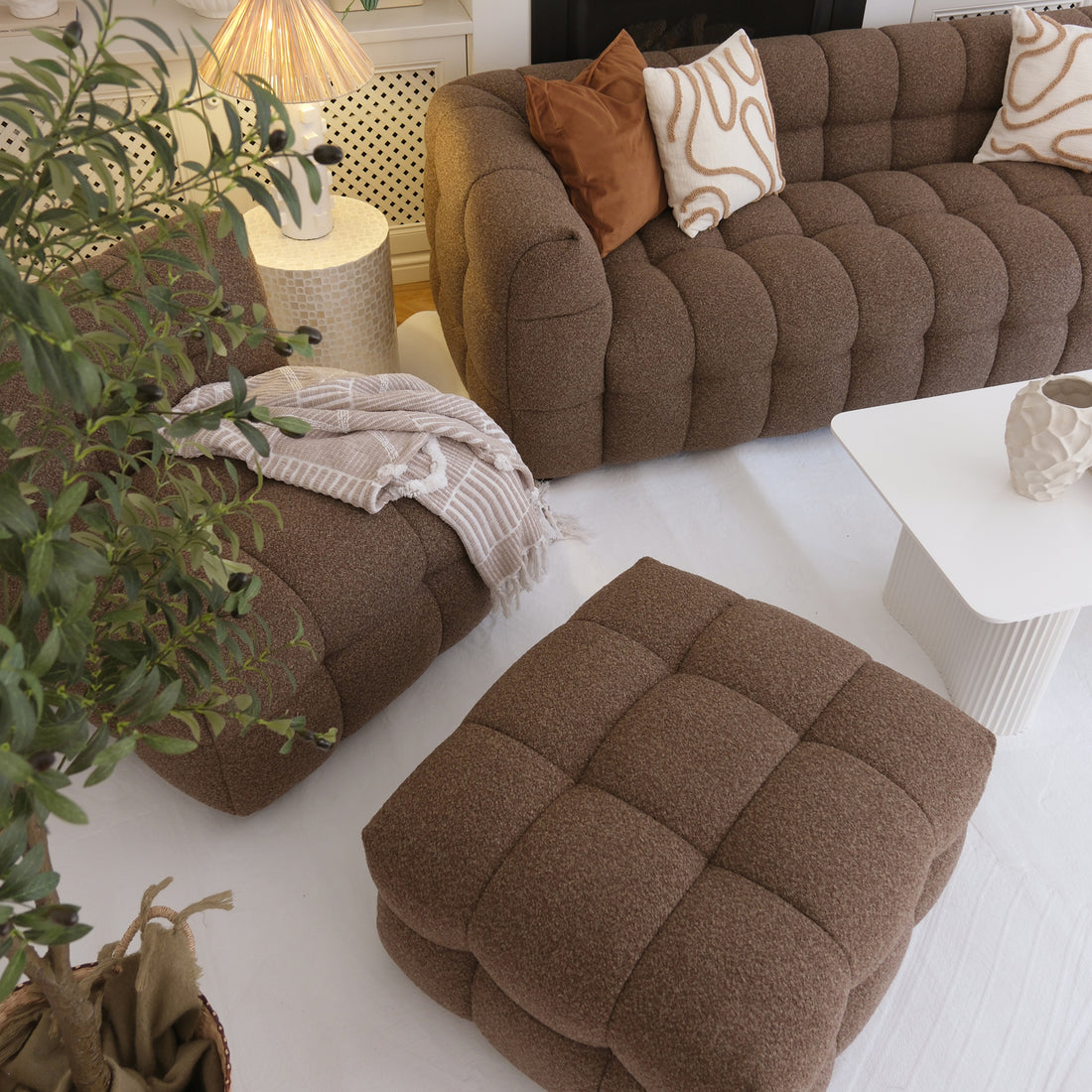 Norah 5 Seater Boucle Sofa Set With Ottoman in Brown, featuring quilted-style texture, rust-brown and cream pillows, atop a pale area rug, accompanied by a white coffee table and leafy plant.