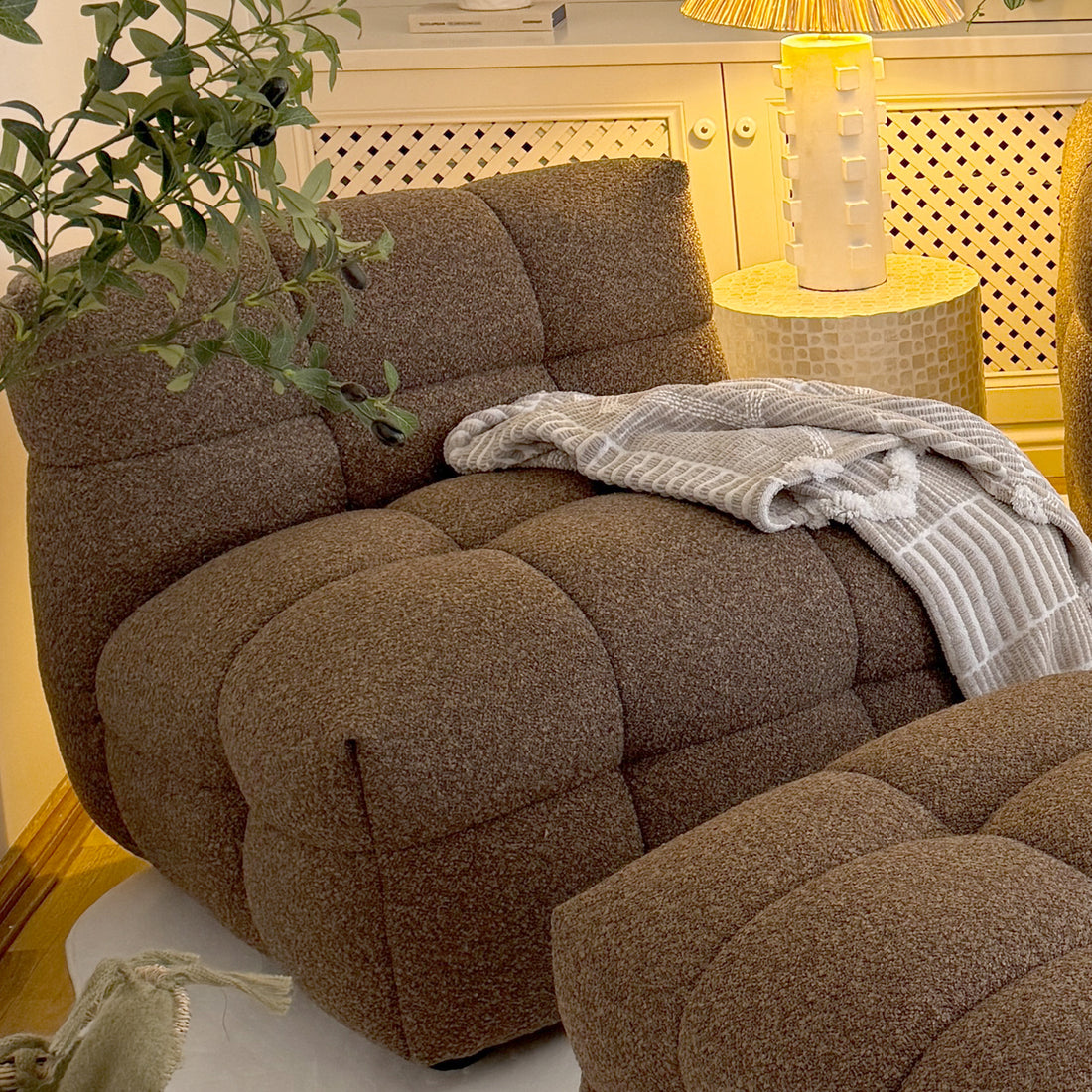 Norah Boucle Single Seater Sofa in Brown, featuring a deep-brown tufted design, is paired with a matching ottoman, accented by a textured throw and adjacent to a mosaic-topped side table.