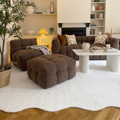 Norah Boucle Ottoman in Brown, featuring a low-profile design with tufted detailing, placed centrally in a cozy living room setup alongside a matching sofa and armchair.