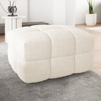 Norah Boucle Ottoman in Cream White, cube-shaped with tufted blocks, upholstered in textured bouclé fabric, sits on a gray rug. Accompanied by a round side table and tall houseplant.